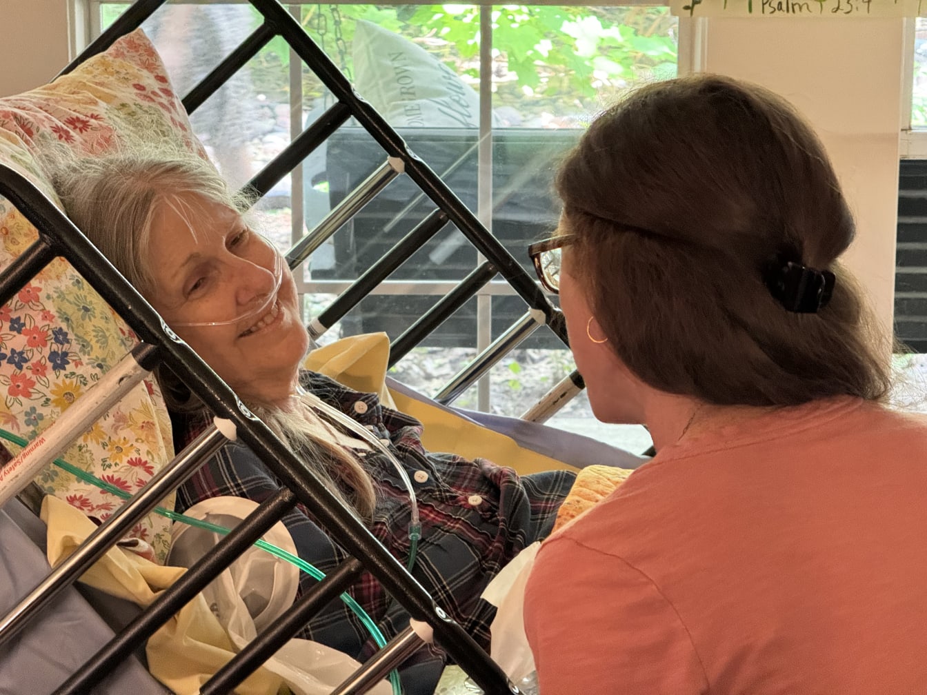 Mom smiling from her hospice bed, on oxygen, as a family member sits beside her by a window