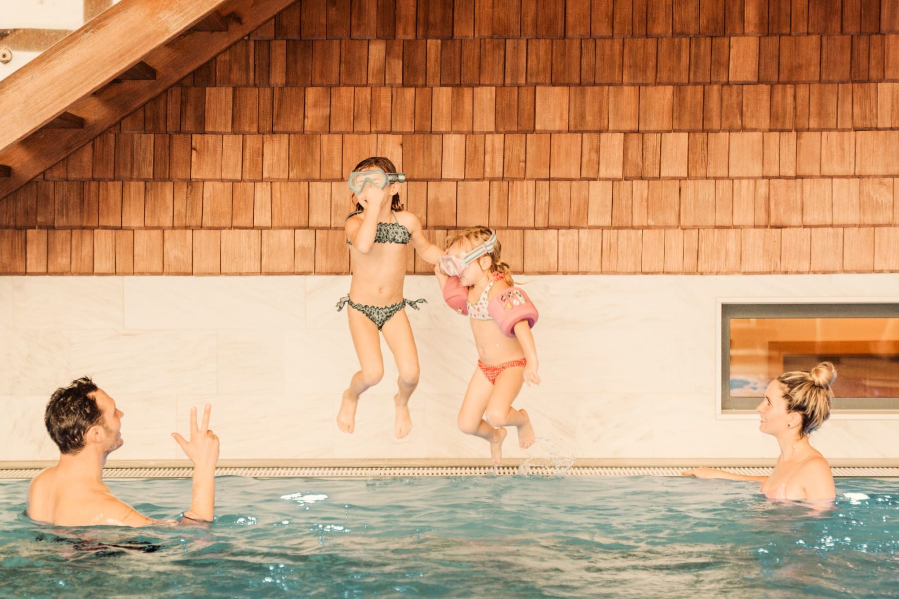 Zwei Mädchen springen im Glemmtalerhof-Pool, während Erwachsene beobachten und Spaß haben.