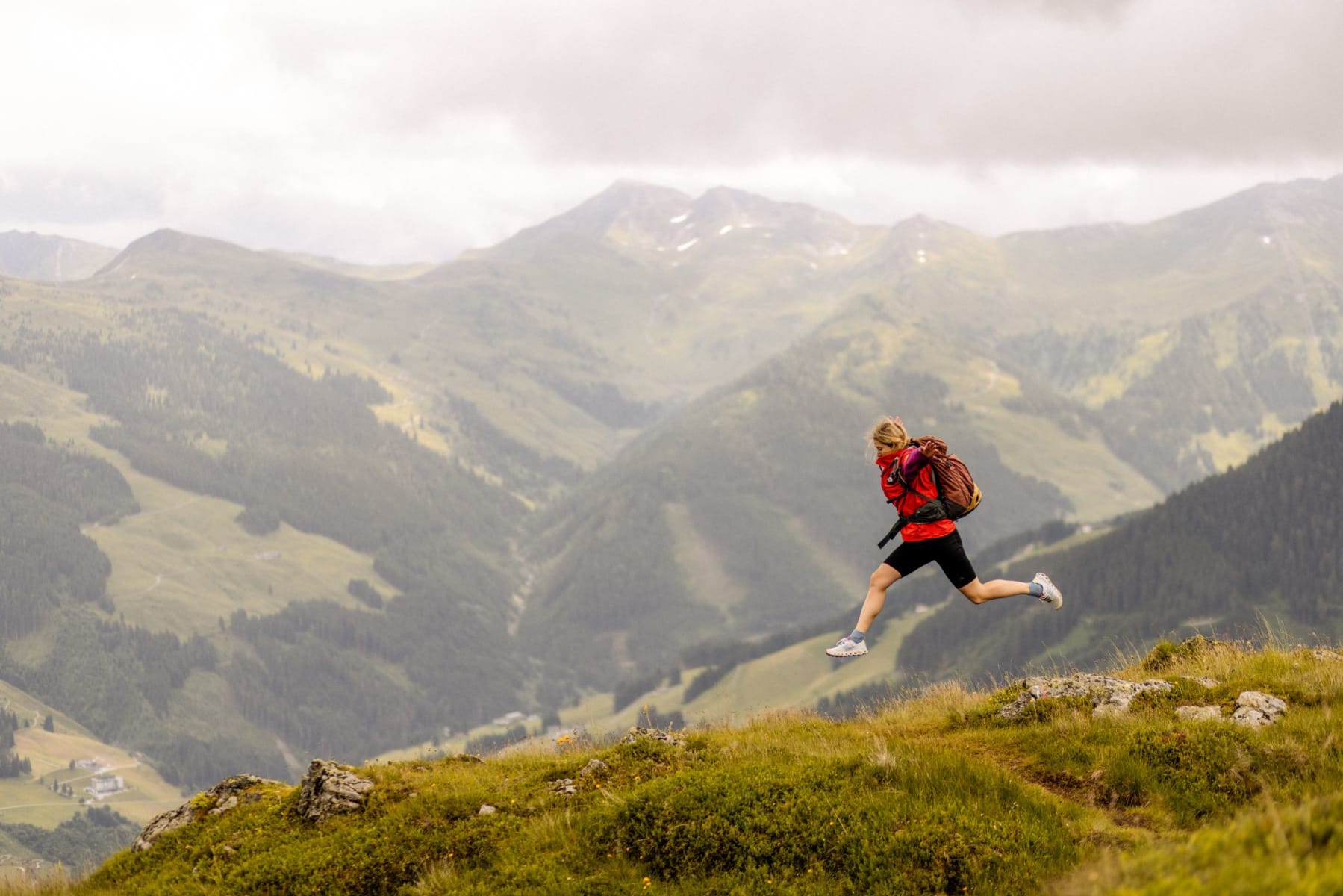 Eine Person springt mit Rucksack über eine Wiese in den Bergen des Glemmtals.
