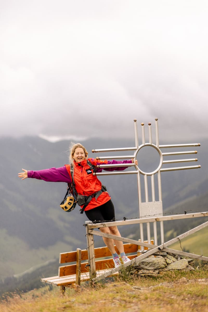 Frau auf einem Gipfel in Hinterglemm, fröhlich vor einem Alpin-Symbol.