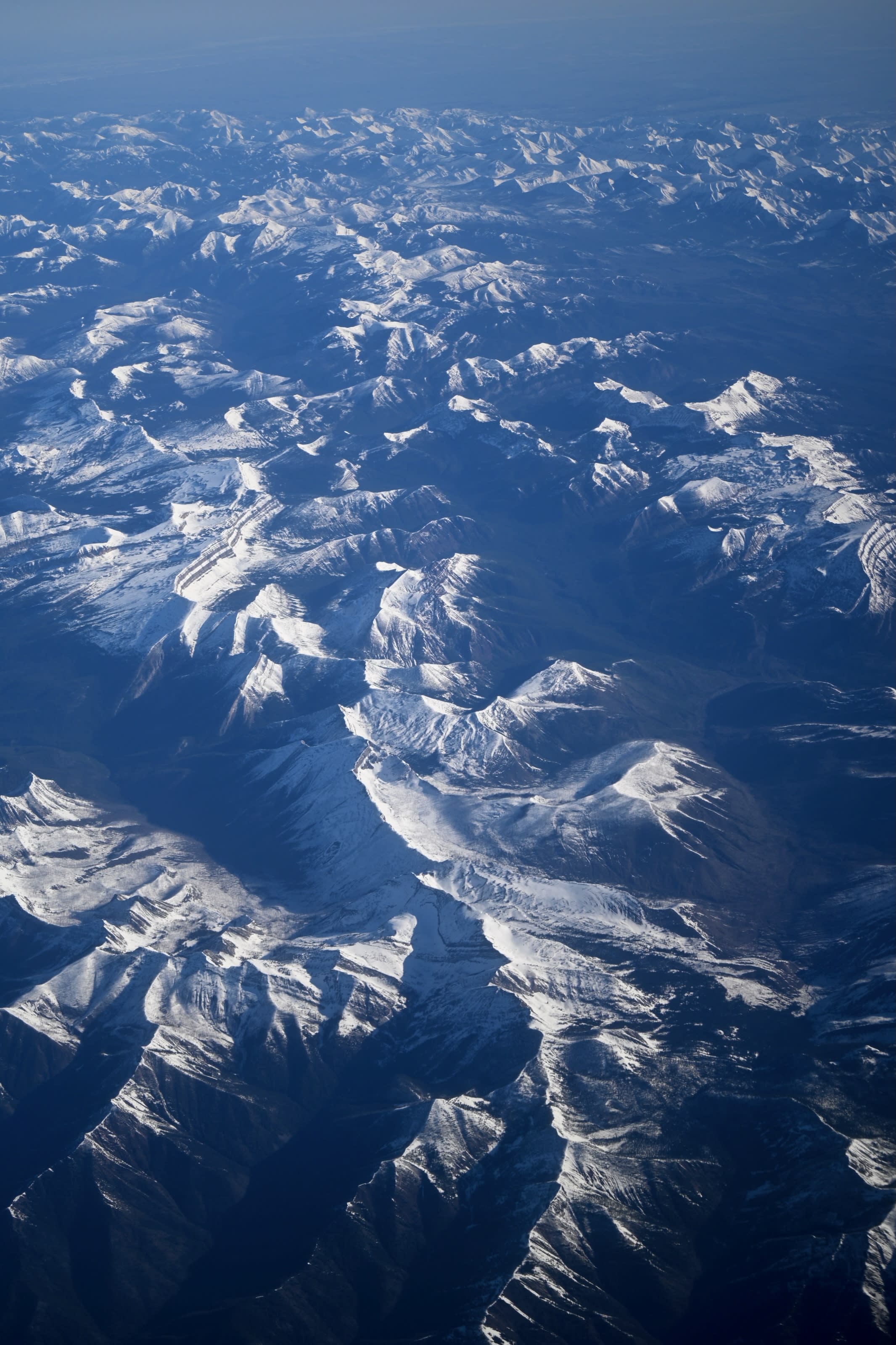 Aerial view of rugged snow-capped mountains stretching into the distance, displaying intricate patterns of peaks and valleys under clear skies.