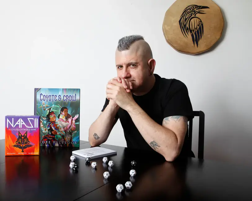 Connor Alexander posa ao lado do livro básico de Coyote & Crow e dados d12 em uma mesa, com um tambor tradicional decorado ao fundo