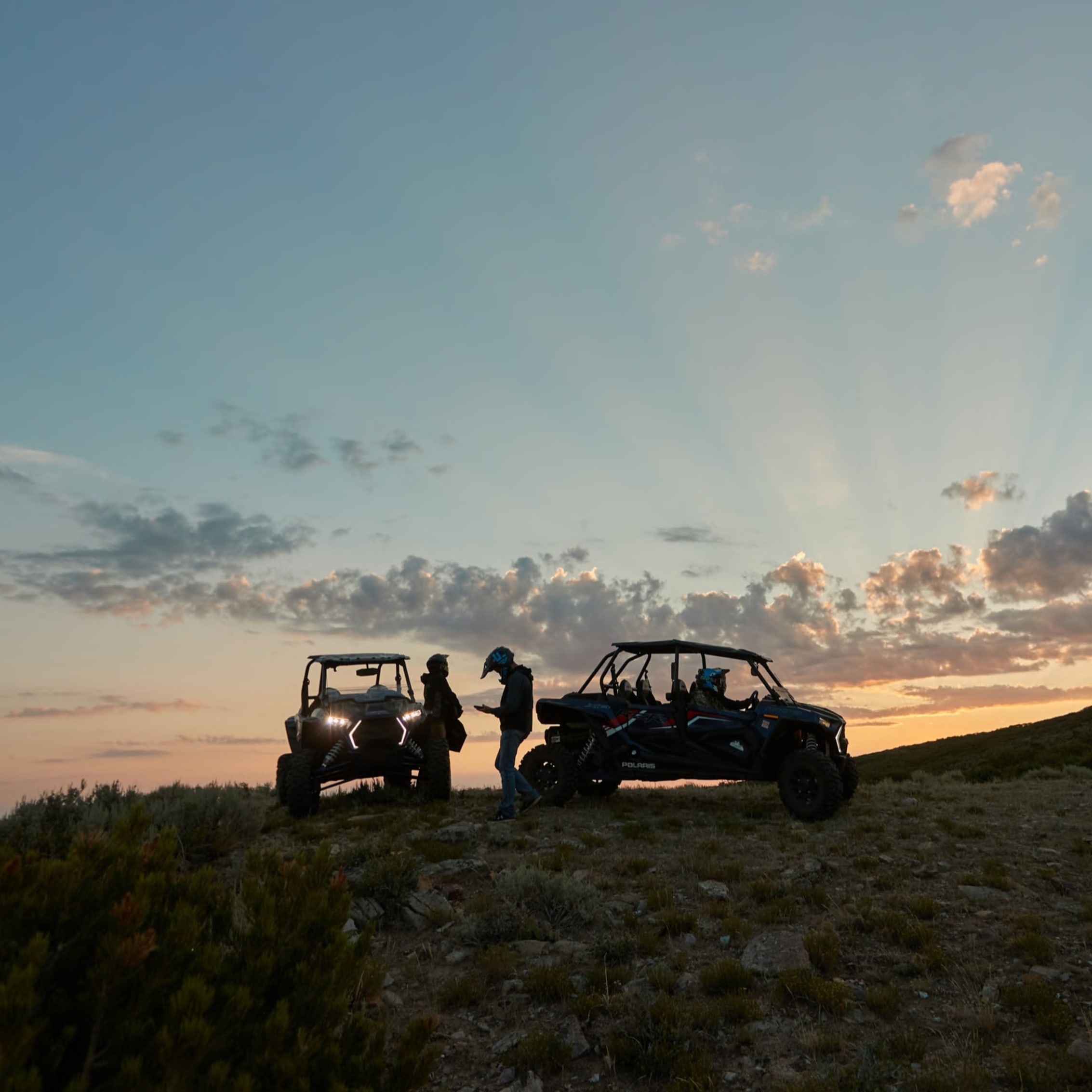 Silhouettes of people and Polaris vehicles on a mountainous horizon at dusk, with a picturesque sky suggesting a peaceful conclusion to a day of self-guided off-road touring.