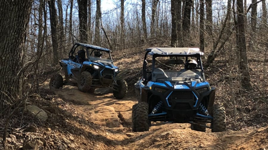 Two Polaris RZR side by sides on dirt wooded trail