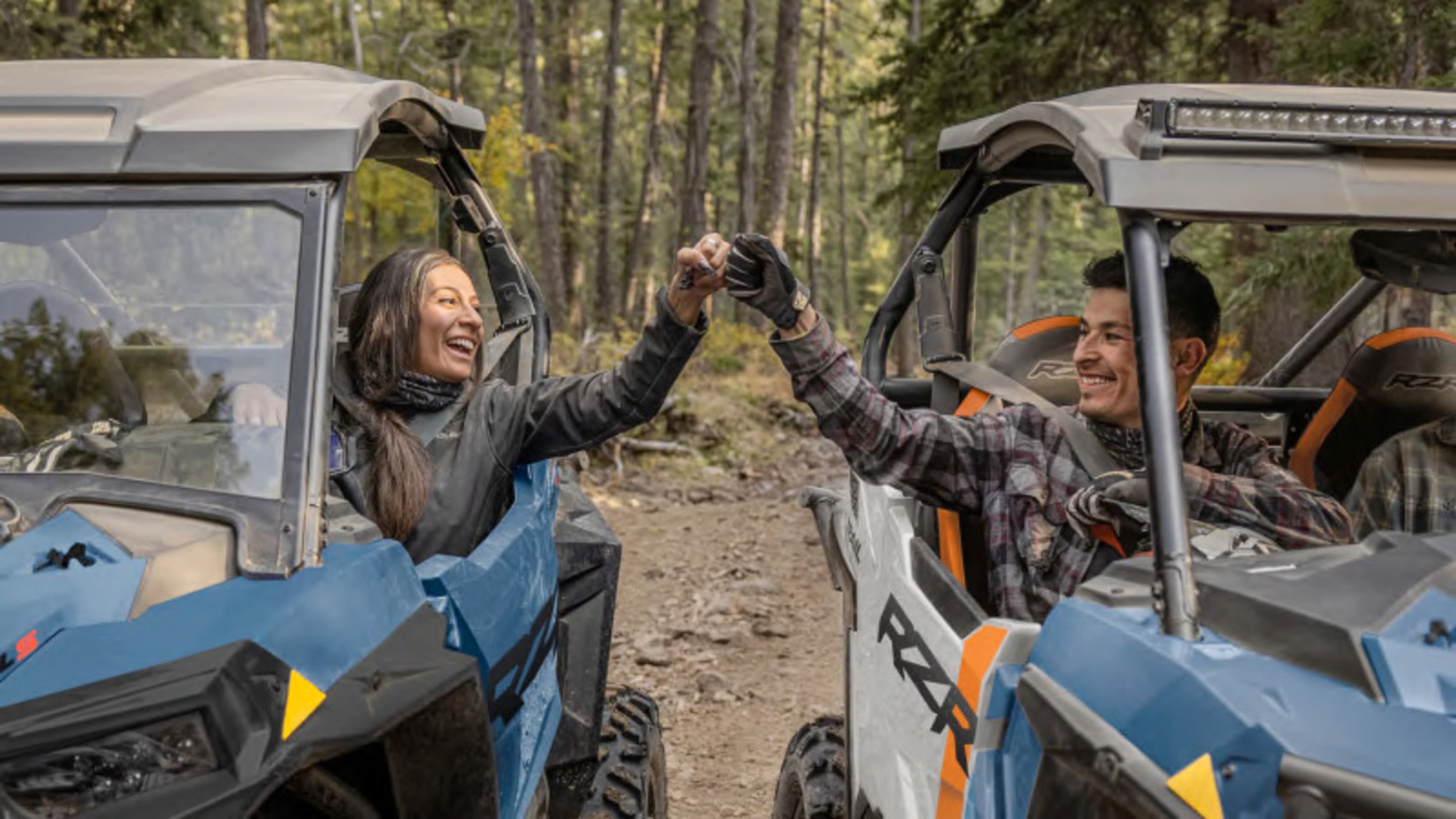 Joyful couple doing a fist bump while seated in Polaris RZR vehicles on a forest trail, signifying teamwork and fun on an off-road excursion.