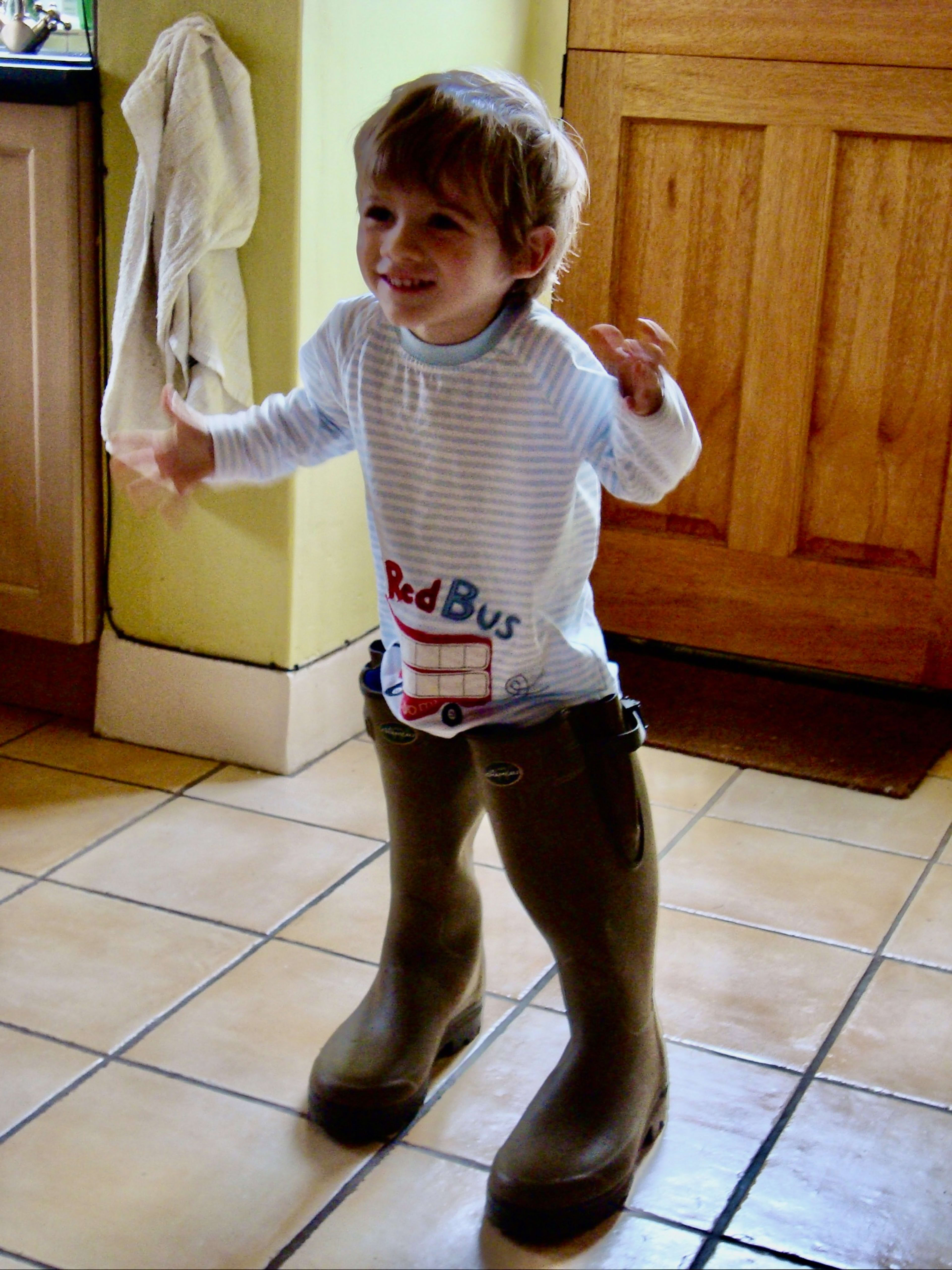 Toddler Charlie in oversized wellies, grinning in the kitchen