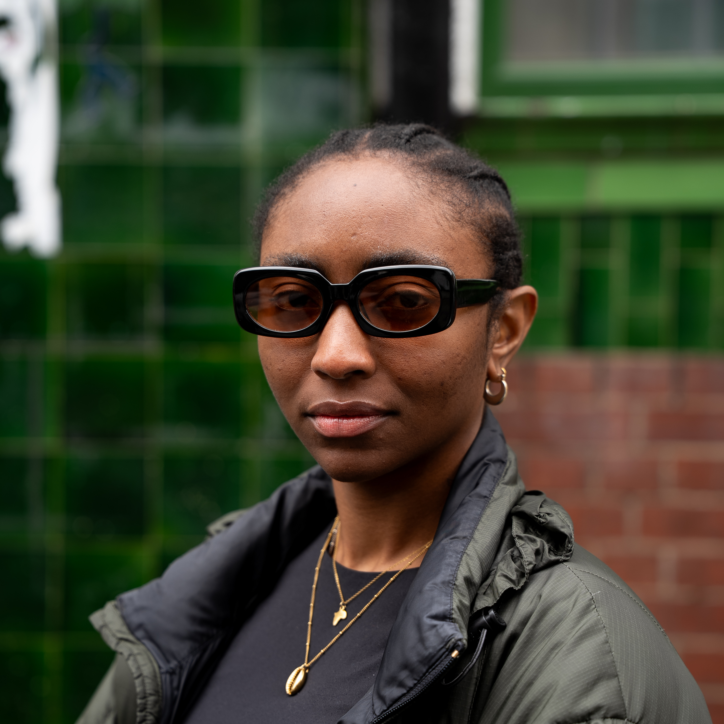 A close up portrait of Esther, she's wearing thick black sunglasses with orange lenses and
             stands infront of a shiney green tiled wall.