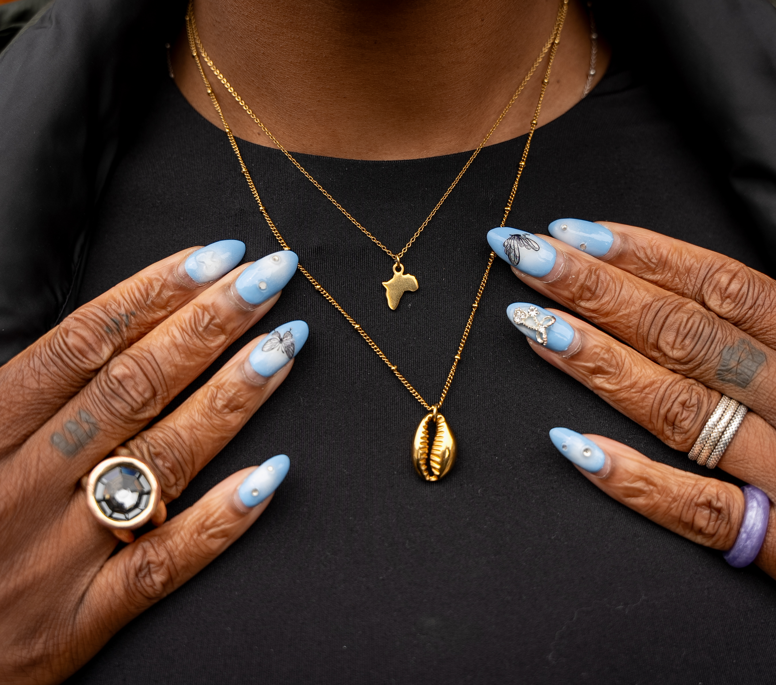 A close up of Esther's hands and neck. She's wearing a thin chain with a pendant in the shape of Africa,
              another longer chain with a gold seashell pendant.
              Her nails are long almond shape in a sky blue colour with 3d chrome designs