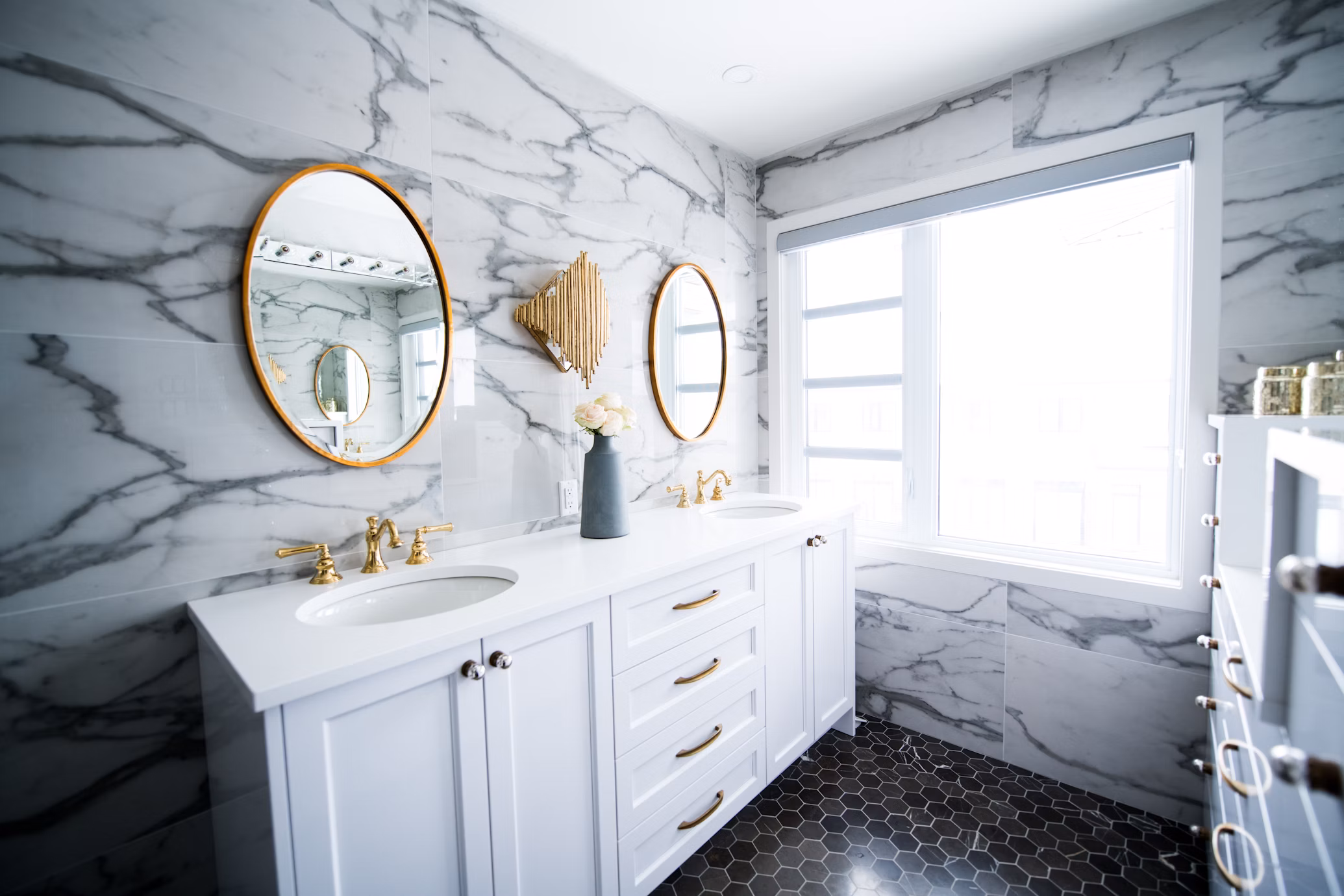 Modern bathroom featuring a porcelain-enamel alcove bathtub framed by rustic wood, with double vanities, vintage wall sconces, and a large window offering natural light—showcasing balance between style, functionality, and durability.