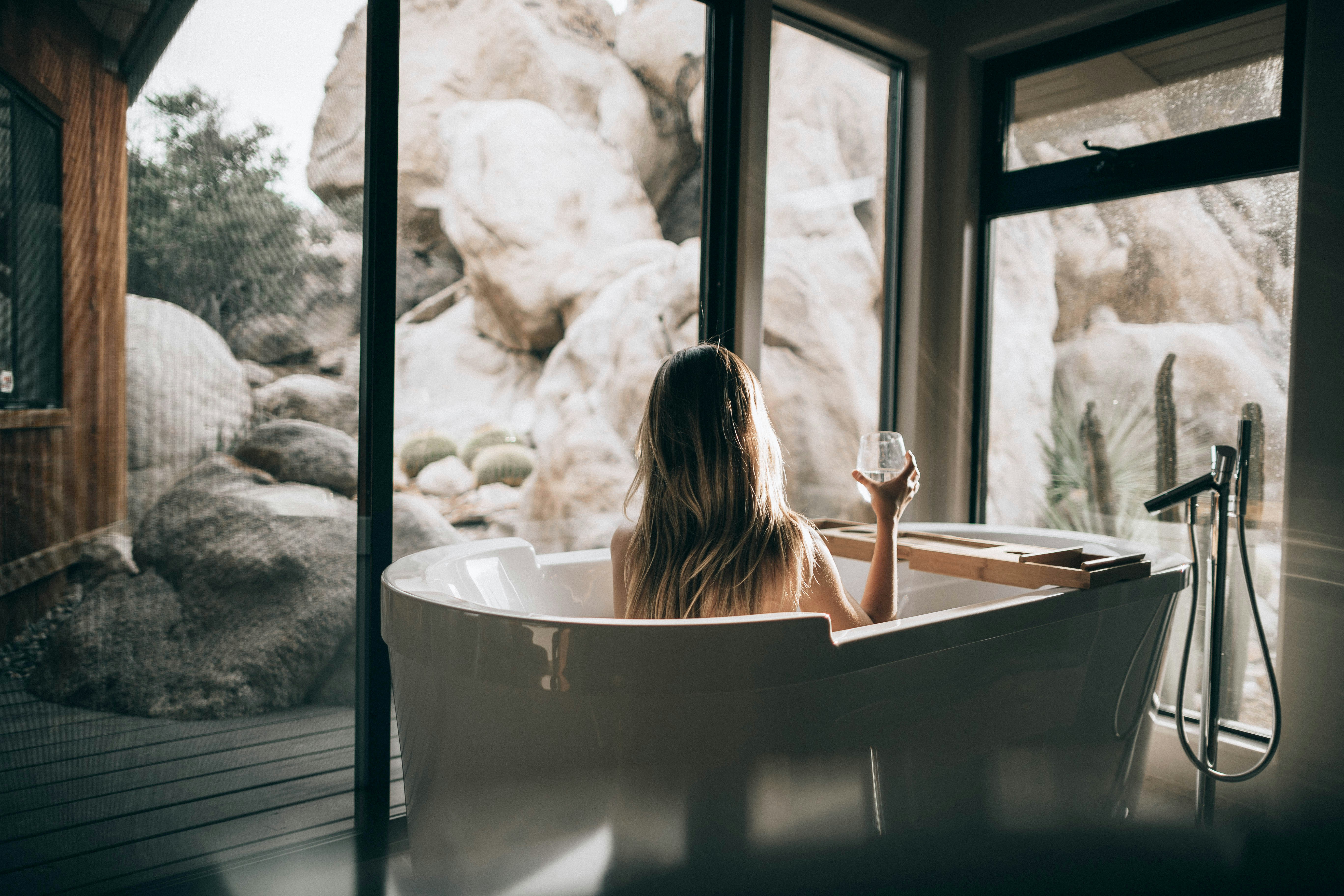 Modern freestanding bathtub with a woman enjoying a glass of wine, surrounded by floor-to-ceiling windows showcasing natural rock formations—creating a luxurious, spa-like bathing experience with relaxation and rejuvenation.