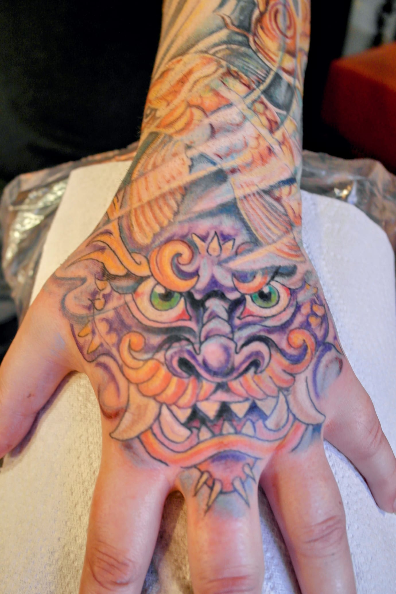 A colorful tattoo of a stylized dragon face with vivid green eyes covers the back of a persons hand. The design features intricate swirls and shading in shades of purple, orange, and blue, extending onto the wrist and arm.