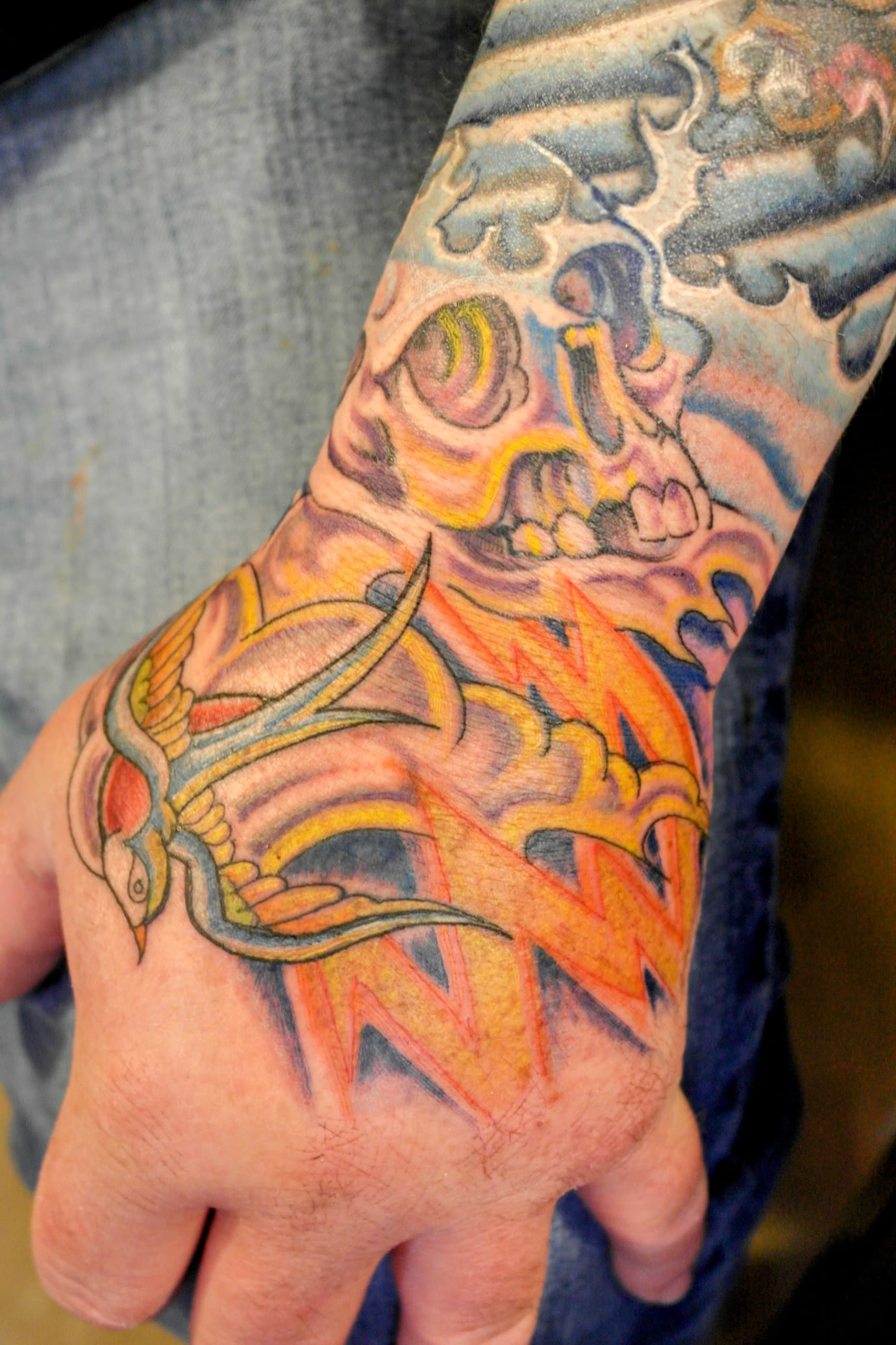 A vibrant tattoo on a hand features a swallow flying amid colorful clouds and lightning bolts. The design incorporates hues of orange, yellow, and blue, extending up the wrist with detailed swirling cloud patterns.