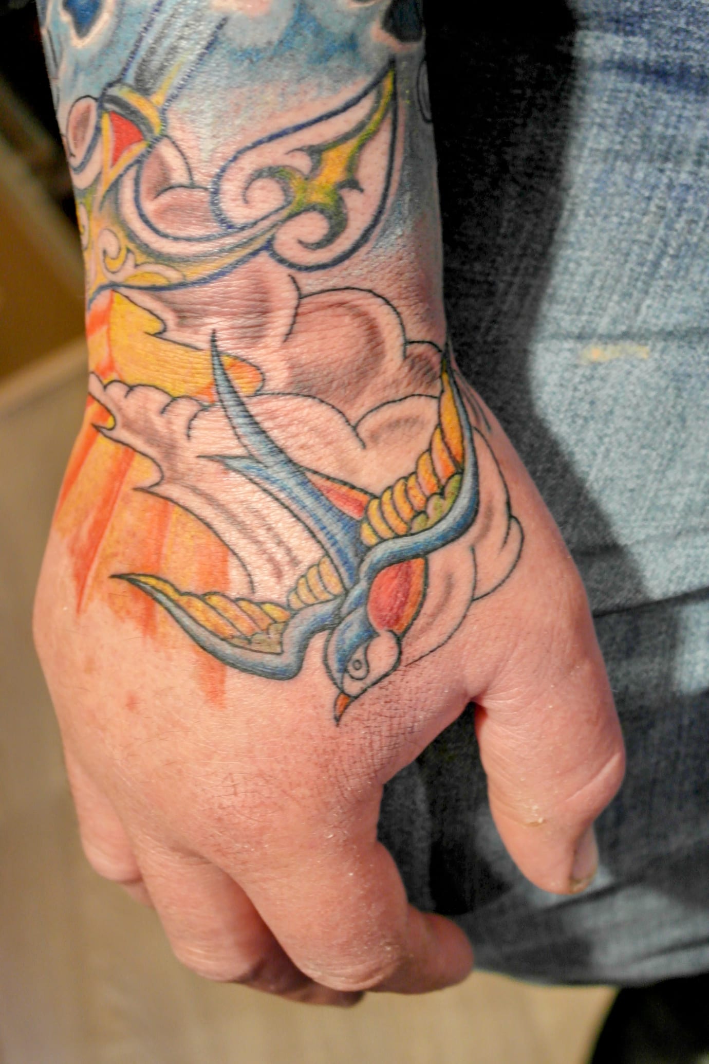 A hand with a colourful tattoo featuring a blue and yellow swallow, surrounded by swirling clouds and a hint of orange and red resembling the sky. The tattoo extends onto the wrist and forearm with intricate patterns.