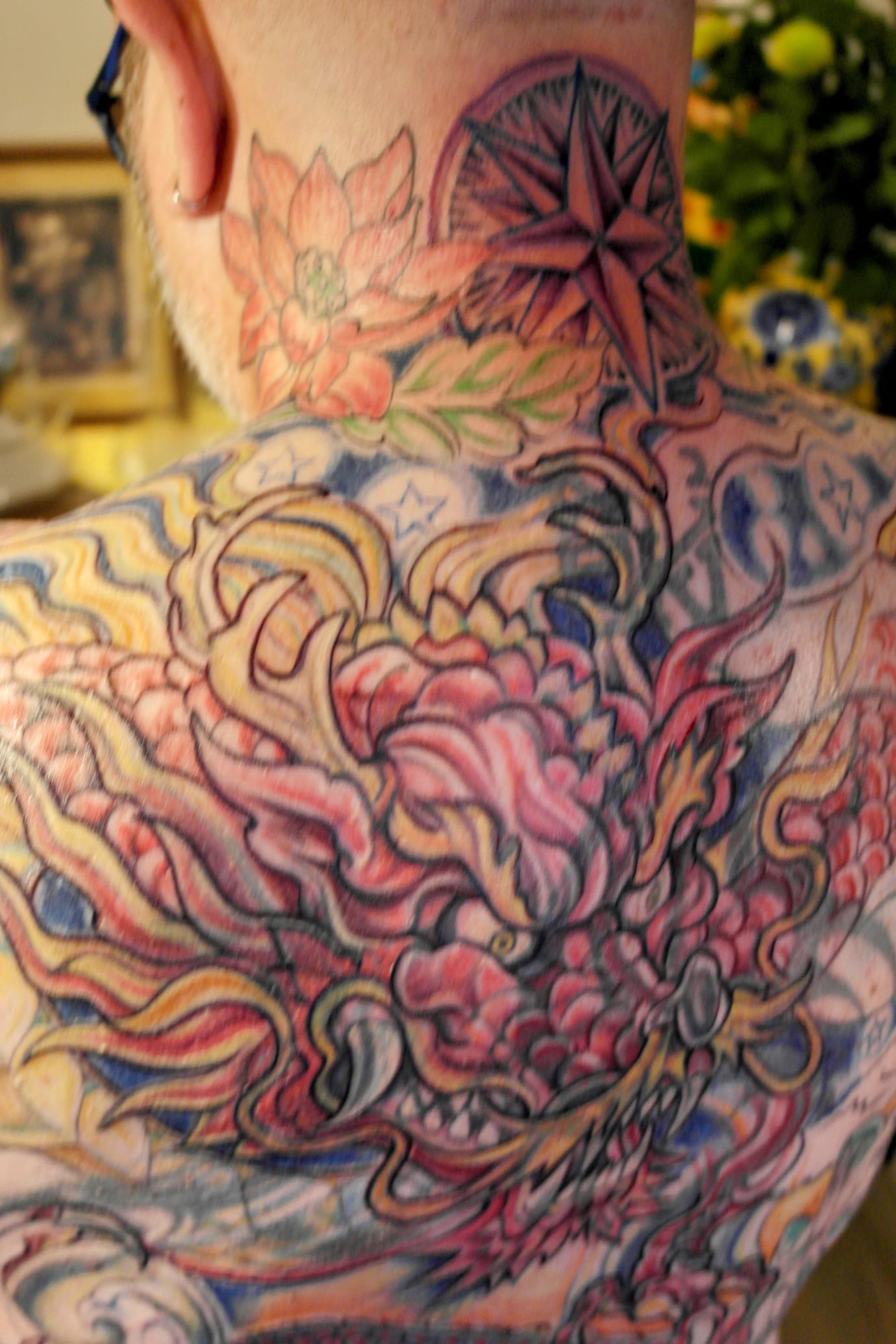 A person with a back tattoo featuring a colorful, intricate design. The tattoo includes a compass rose, floral elements, and swirling patterns. The image is focused on the back, revealing detailed, vibrant artwork.