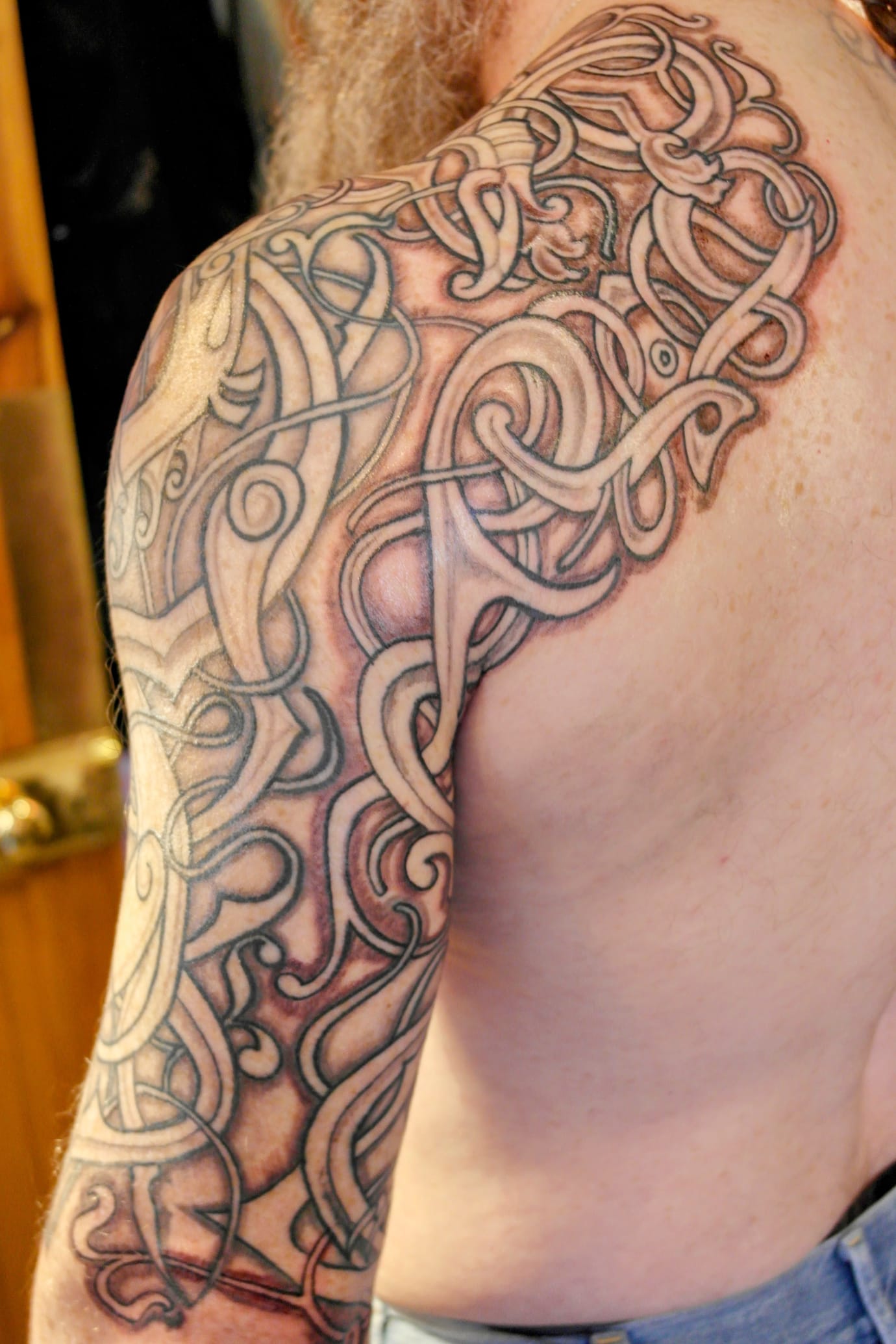 A person with a large, intricate Celtic-inspired tattoo covering their shoulder and upper arm. The design features interwoven patterns with shading and detailed linework.