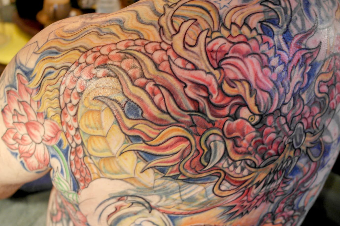 Back tattoo featuring a colourful, intricate design of a dragon with scales, claws, and swirling patterns. The dragon is surrounded by floral elements, including a large flower on the shoulder, all in vibrant shades of red, yellow, and blue.