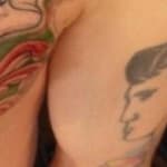 Close-up of a colorful chest tattoo featuring a large winged design in shades of orange, yellow, and green. The words Rock & Roll are prominently inked in a classic script. Framed pictures are blurred in the background.