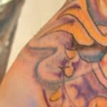 A colorful tattoo of a stylized dragon face with vivid green eyes covers the back of a persons hand. The design features intricate swirls and shading in shades of purple, orange, and blue, extending onto the wrist and arm.