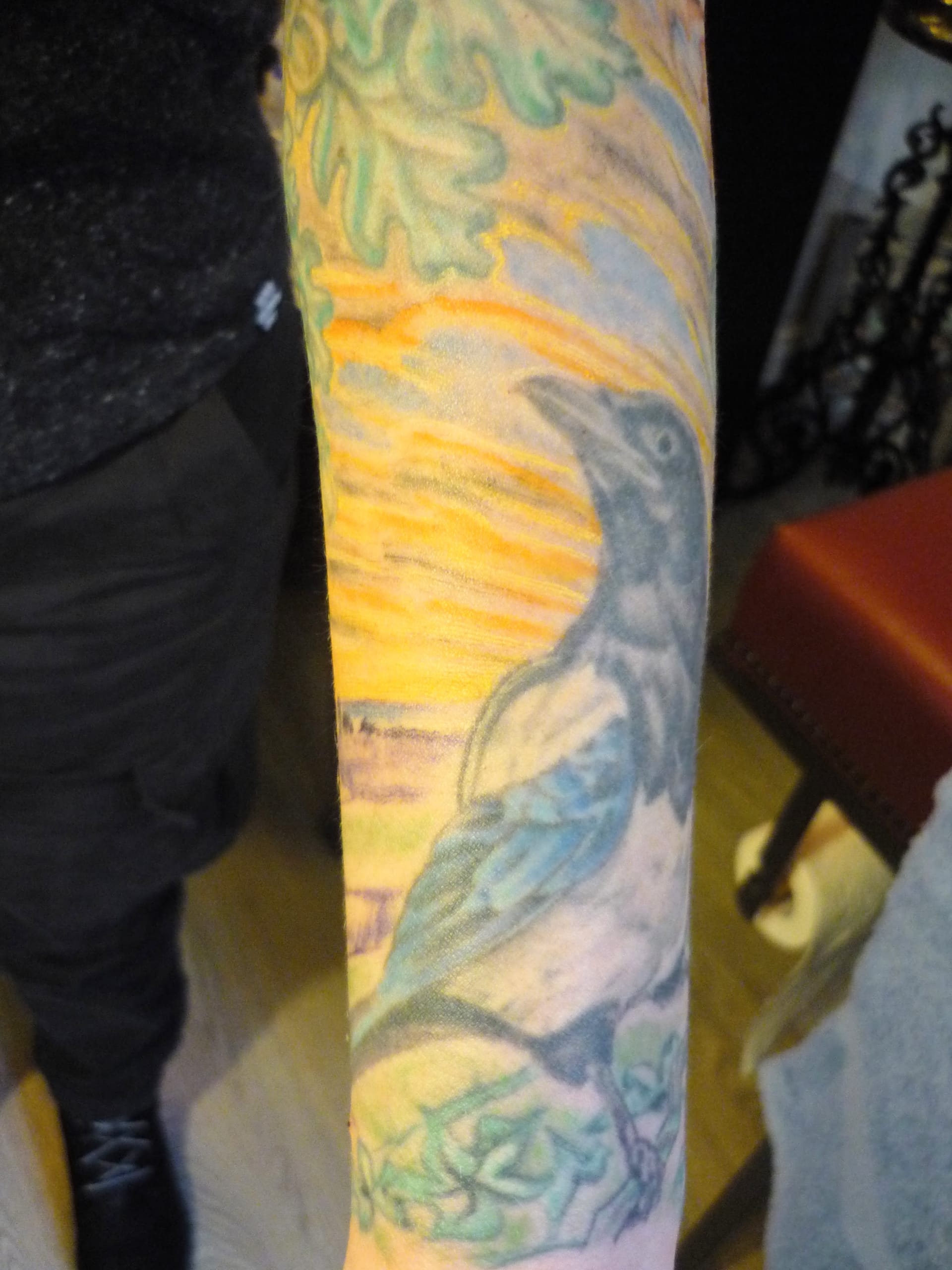 Forearm tattoo featuring a detailed crow surrounded by green leaves against a vibrant yellow and orange background.