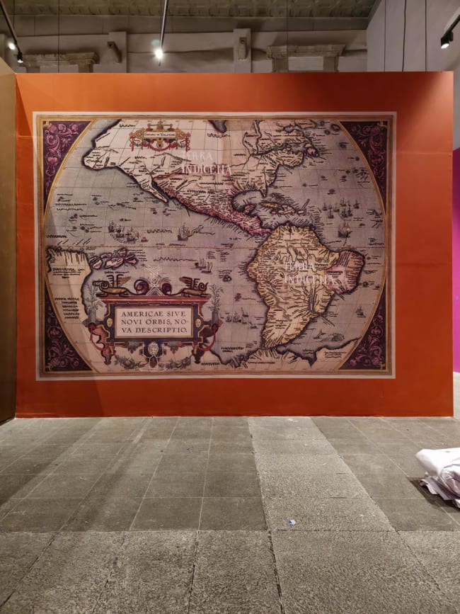 Pieza cartográfica de gran formato enmarcada y montada sobre muro naranja en sala de exposición