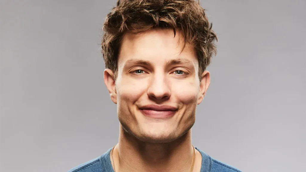 Matt Rife smiling in a close-up portrait wearing a blue shirt, photographed against a plain gray background.
