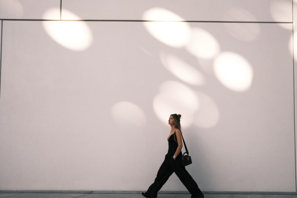 a woman walking down a sidewalk past a white wall