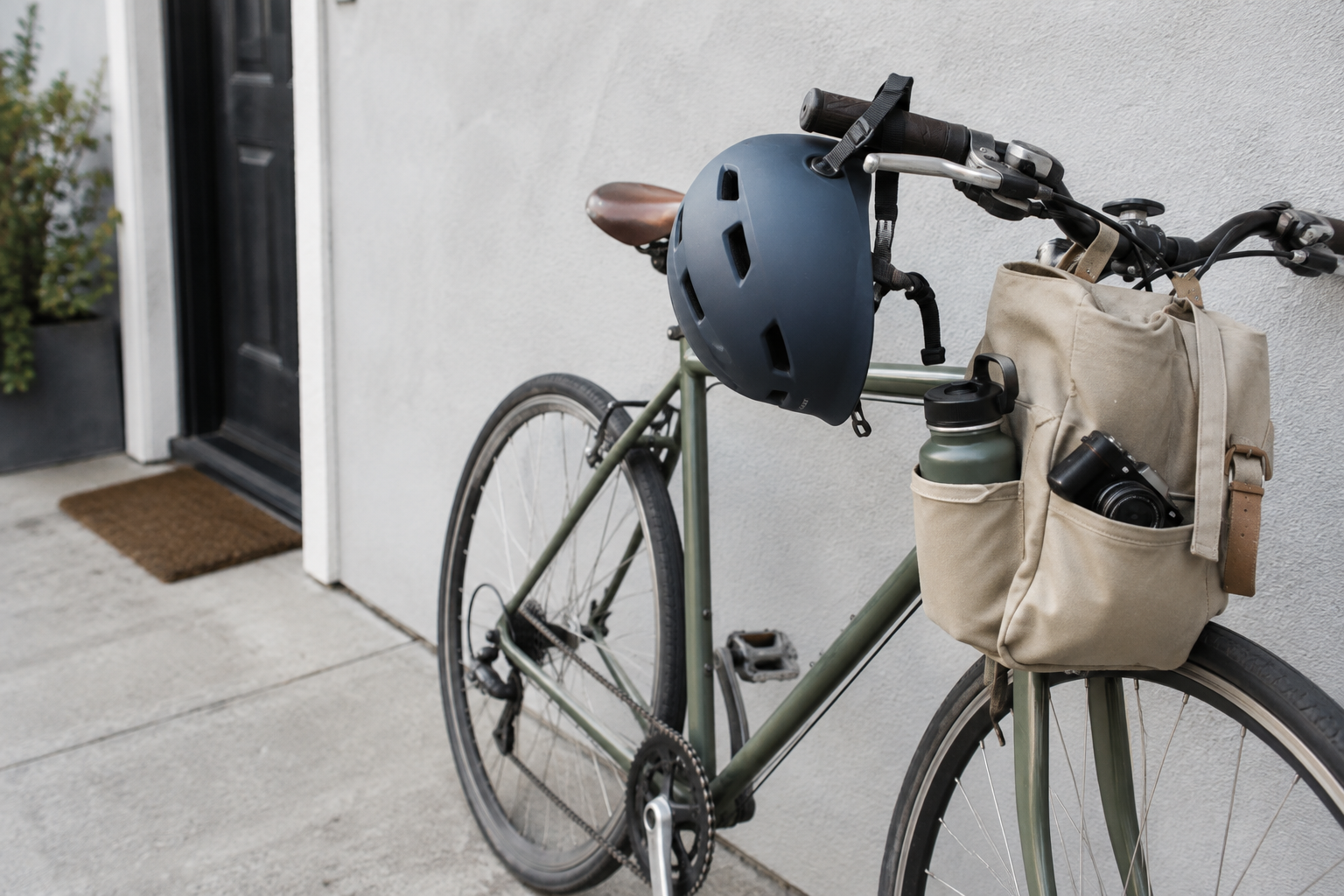 A bicycle, helmet and day bag with a water bottle and camera beside an exterior wall.