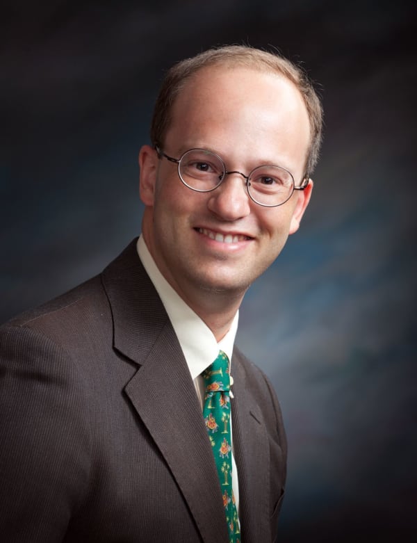 Ritratto di Daniele Camiz n giacca marrone e cravatta verde su sfondo sfumato da studio.