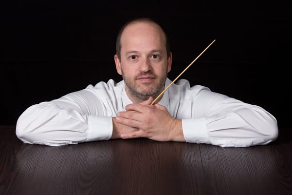 Ritratto si Daniele Camiz con camicia bianca, braccia sul tavolo e bacchetta fra le mani.