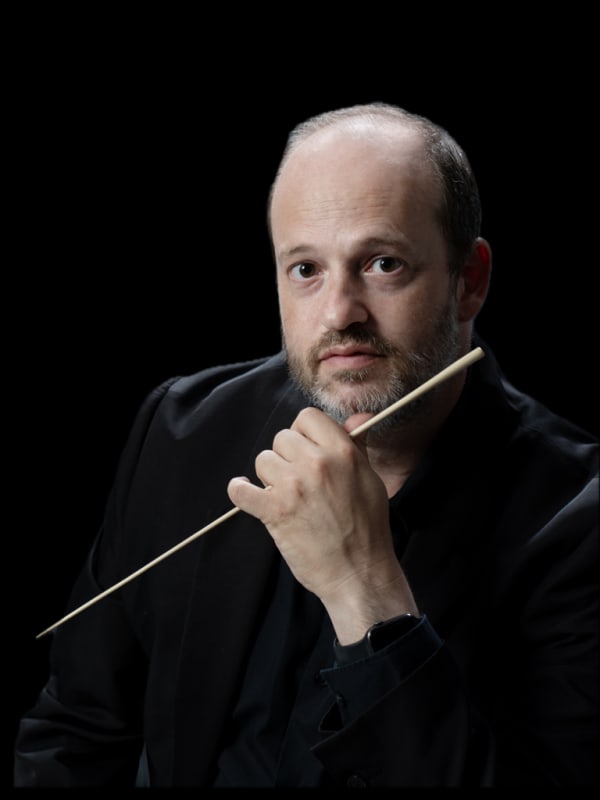 Daniele Camiz seduto, con la bacchetta appoggiata al mento e lo sguardo in macchina, su sfondo nero.