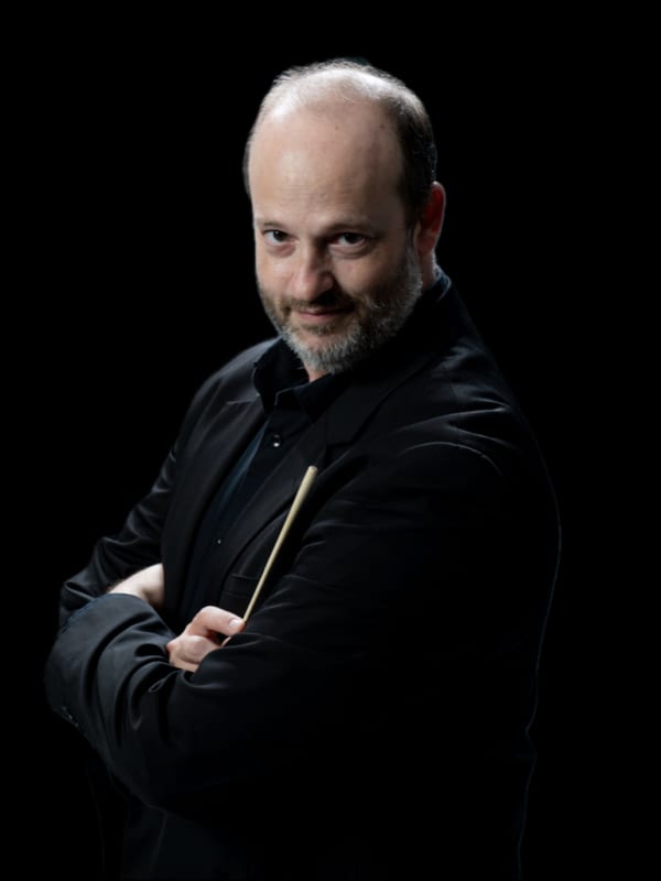 Daniele Camiz con braccia incrociate, vestito di nero, con bacchetta in mano su sfondo nero.