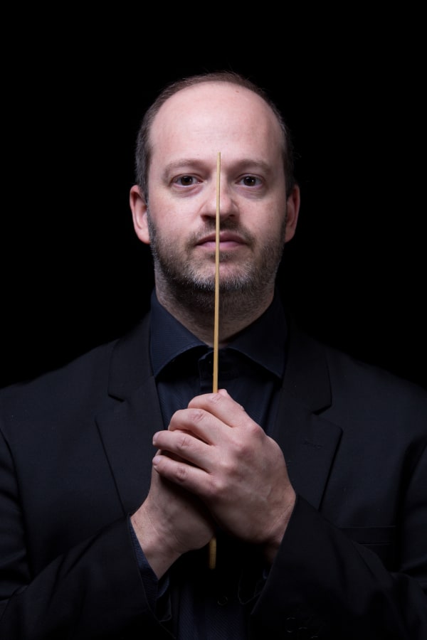 Ritratto frontale con bacchetta tenuta al centro del volto su sfondo nero.