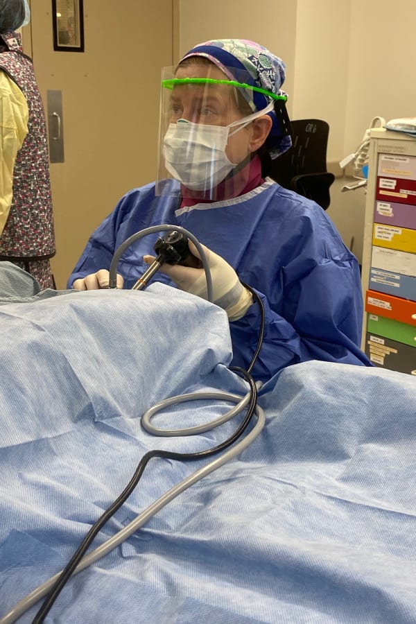 Doctor wearing a face shield with a 3D printed visor and surgery garments holding a laparoscope in an operating room, performing surgery on a patient covered in blue fabric.