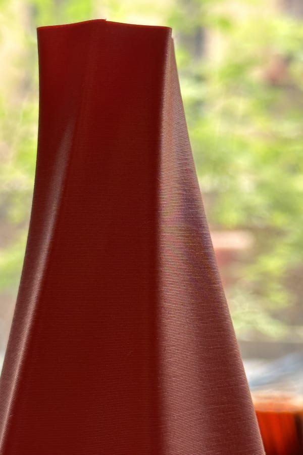 Close view of a terra cotta-colored vase top with a gently twisting rippled surface, showing layers highlighted by the side lighting. Behind the vase is out-of-focus greenery.