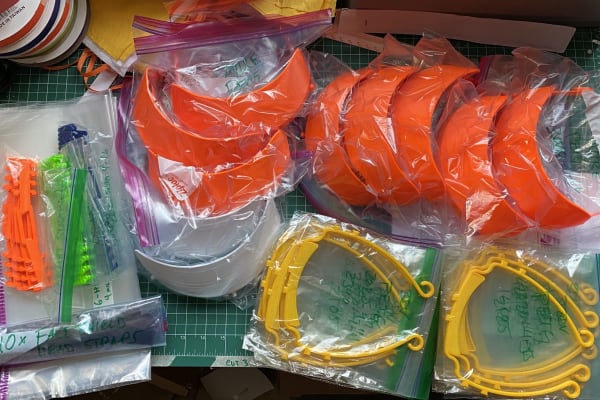 3D printed face shield visors and ear savers packaged in plastic baggies, arranged on a work surface. The bags are labeled in permanent marker with their packing dates and material usage.