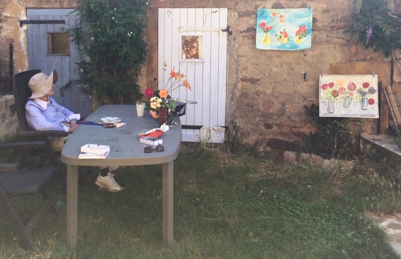 Elderly woman with gray hair sitting in a sunlit garden, looking thoughtful.