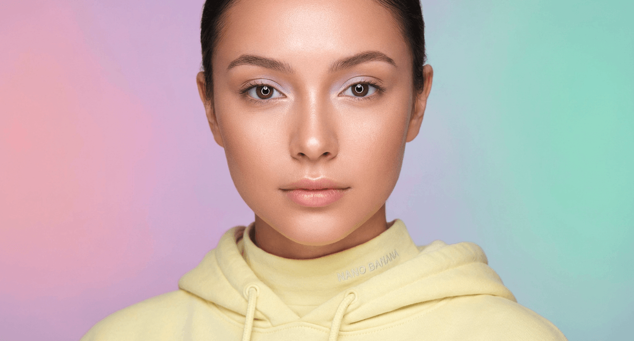 Close-up beauty portrait of a woman with flawless skin and subtle makeup against a soft purple-to-mint gradient background, wearing a pale yellow hoodie with "NANO BANANA" text.