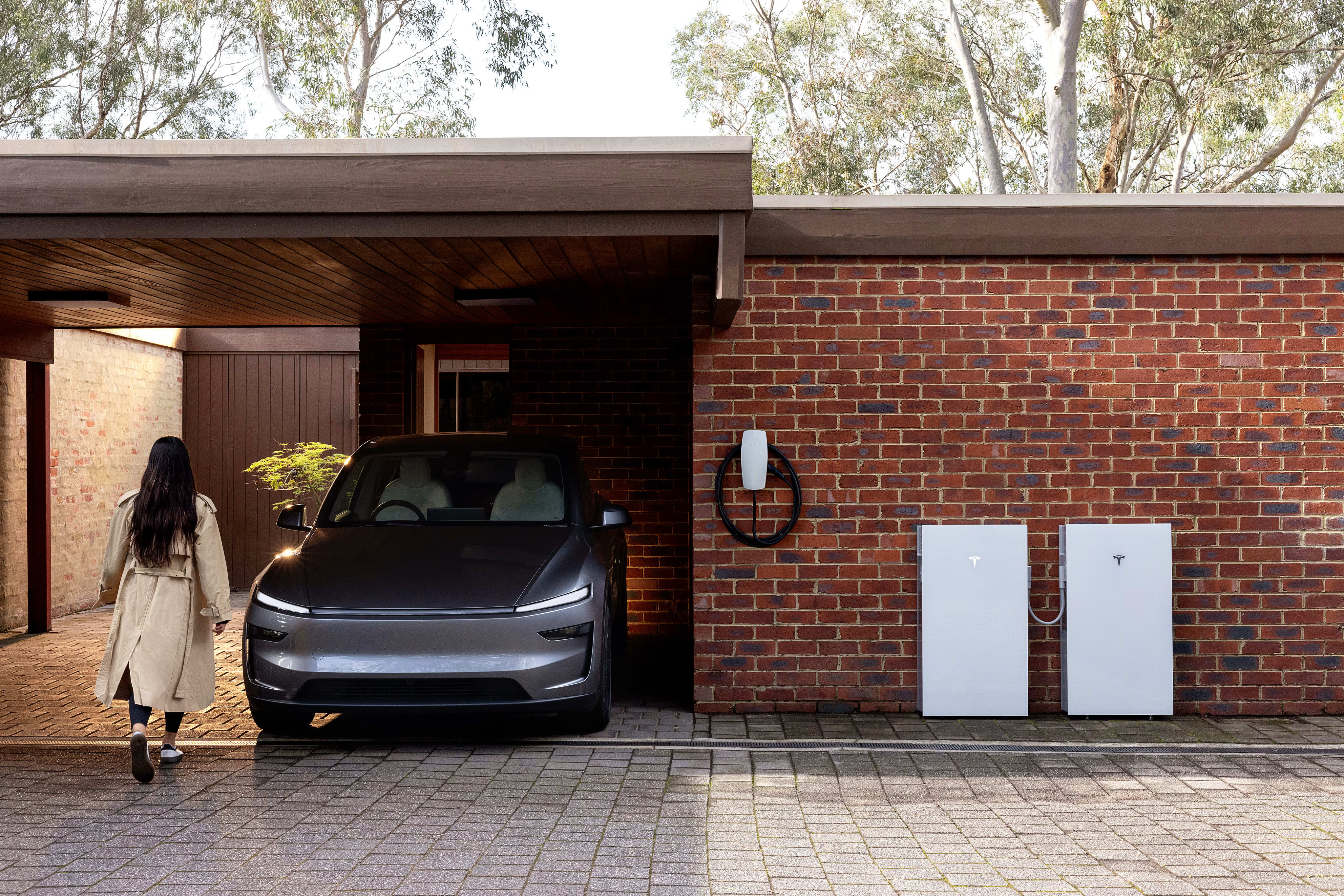 Two Powerwall units and a Wall Connector sit in front of a home's exterior brick wall as a woman walks past a Model Y 