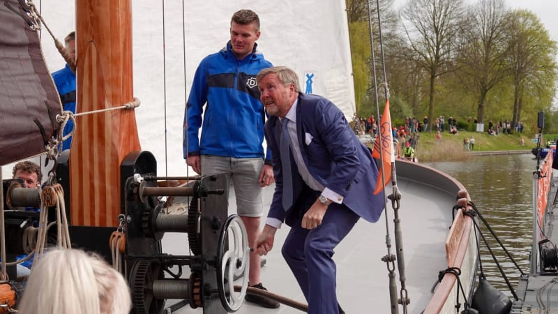 Een man in pak buigt voorover bij het ankermechanisme van een historisch zeilschip terwijl een jongere man in blauw jasje toekijkt.