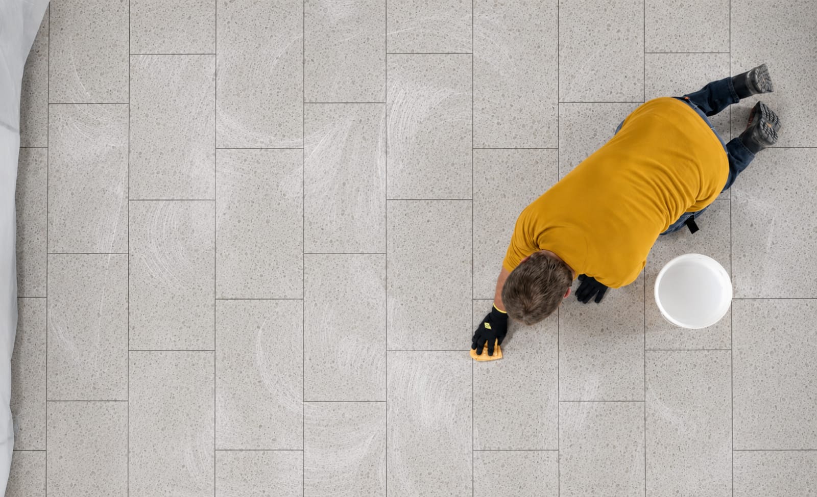 Overhead view of a tiler wiping grout haze from a pale terrazzo-look tiled floor in an Australian home, with a sponge and bucket.