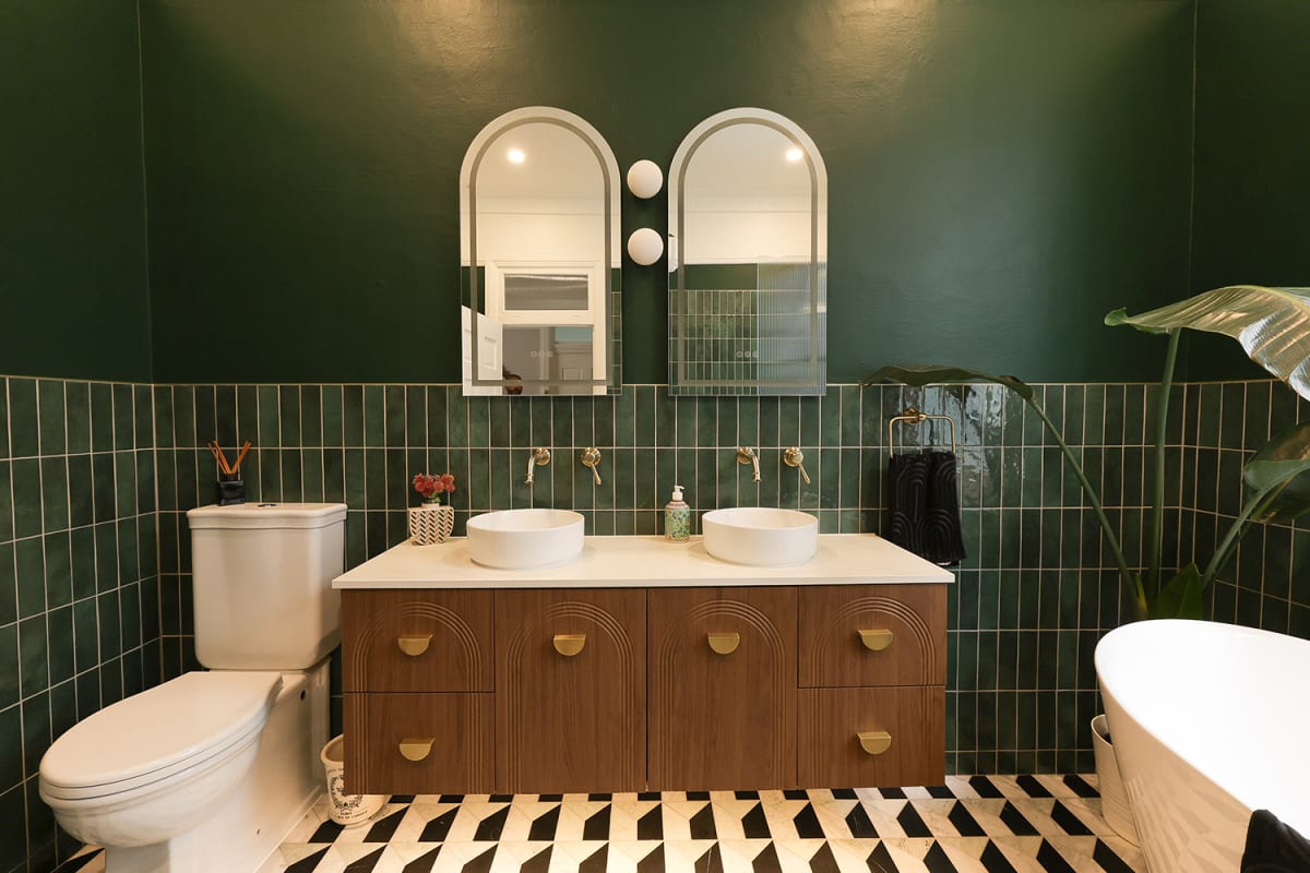 A Sydney bathroom featuring a deep forest green handmade Moroccan zellige feature wall laid in vertical stack bond, with twin arched mirrors and white globe sconces above a dark walnut fluted vanity with brass half-moon handles, white vessel basins on a stone benchtop, brushed brass wall-mounted tapware, and a dramatic black-white-and-grey geometric cubed marble floor. Zellige used as a confident colour statement in a moody, design-led Sydney ensuite.