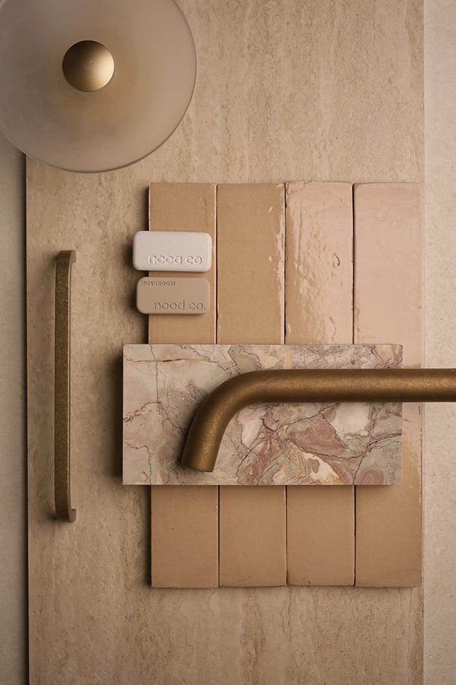 A palette flatlay on a travertine surface — Zellige Look Feature Wall Tile in natural, an Italian-made ceramic look-alike laid in slim 50×150mm vertical format with the soft hand-touched glaze and natural variation of real zellige. Paired with a brushed brass linear cabinet handle, a smoked glass dome wall sconce with brass ball, a brushed brass wall-mounted basin spout, two Nood Co. bathware sample chips in cream and mushroom, and a sage-and-blush veined marble vanity sample. A complete palette curated by the Design Tiles team at our Rockdale showroom.