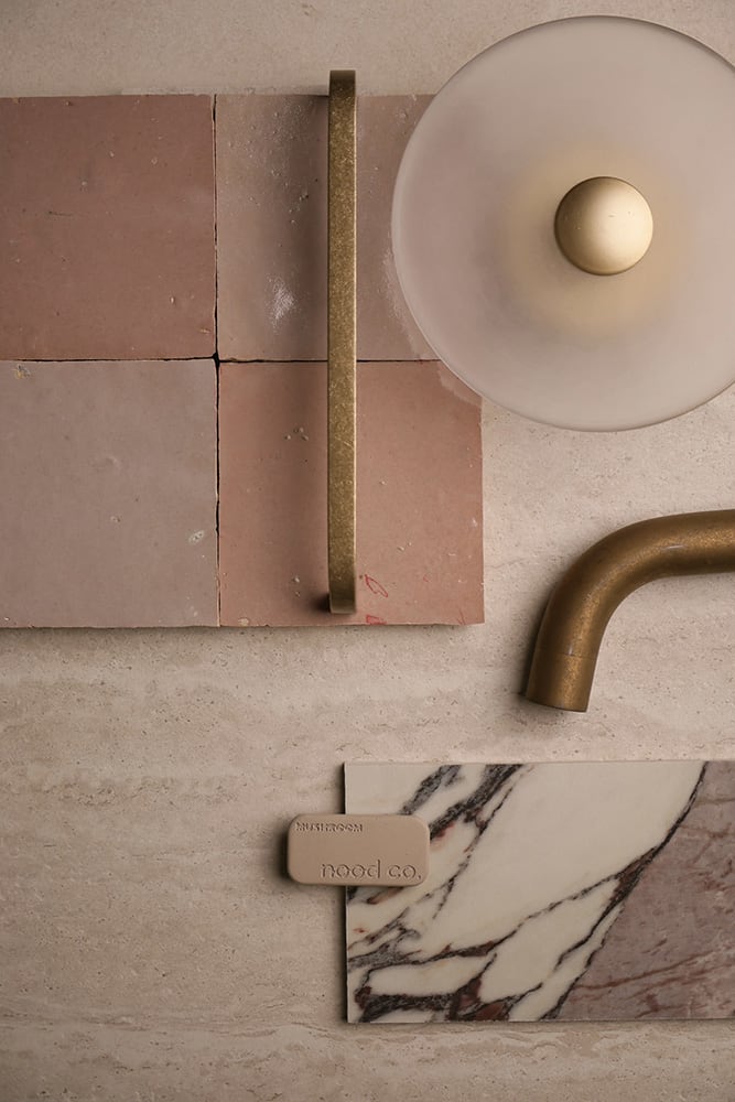 A second palette flatlay on travertine — Zellige Moroccan Tile Handmade Glazed in blush, authentic Fez-made zellige with raw clay edges and hand-poured glaze variation. Paired with a brushed brass cabinet handle, a smoked glass dome wall sconce, a brushed brass basin spout, a Nood Co. mushroom-toned bathware sample, and a Calacatta Viola marble vanity sample with dramatic burgundy veining. A second palette direction curated by the Design Tiles team at our Rockdale showroom.