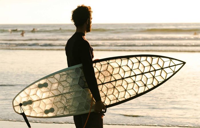A surfer carrying a WYVE 3D-printed eco-surfboard on the beach at golden hour