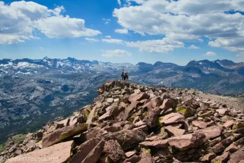 Two hikers on a very rocky mountain top with distant mountains, lakes, and clouds in the sky