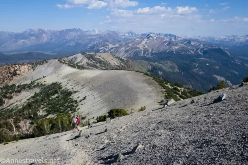 Two hikers on a trail on a gravel mountainside with a mountain ridge and distant mountain peaks