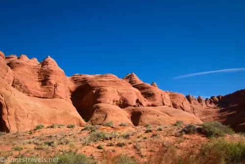Red slickrock cliffs topped with domes above sandy desert and below blue skies