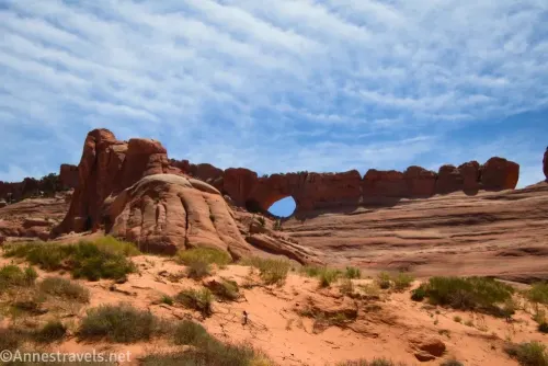Clouds over a slickrock fin with an arch in it above a sandy hill with bushes on it