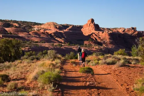 Two hikers on a dirt road bordered by desert brush with red rock formations and cliffs beyond