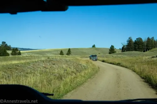 A truck on a dirt road between grassy meadows and a few trees 